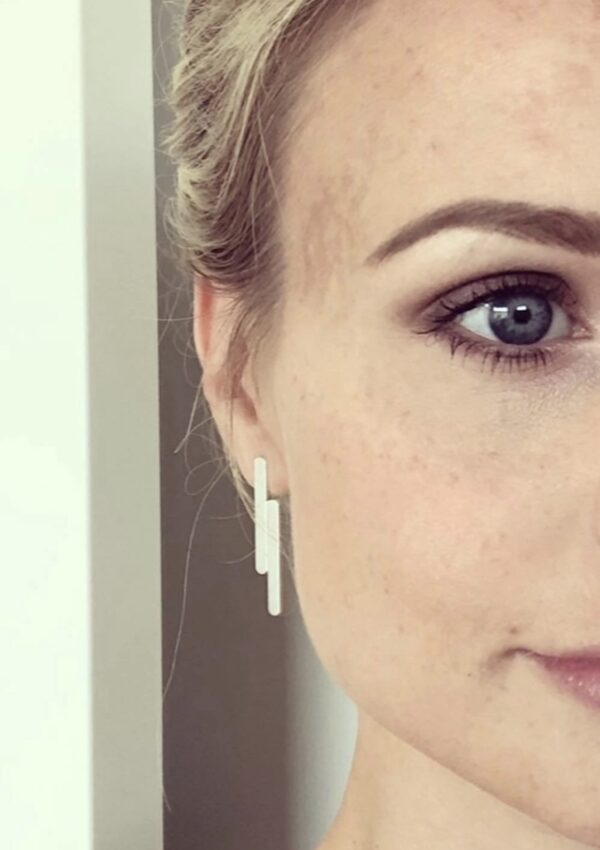 A half of a female face wearing silver earrings shaped as two flat sticks unsymetrically attached to each other.