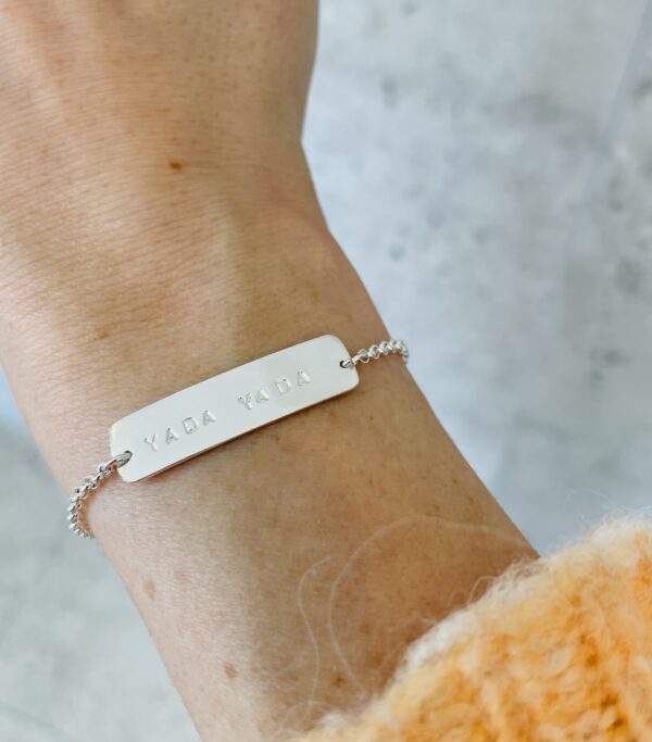 A wrist wearing a silver bracelet with the words "Yada Yada" on it